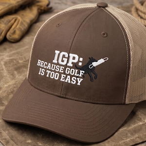 May include: A brown and tan trucker hat featuring an embroidered design of a black dog holding a white object in its mouth. The text reads: IGP: BECAUSE GOLF IS TOO EASY. The hat has a solid brown front panel and a tan mesh back with a button on top.