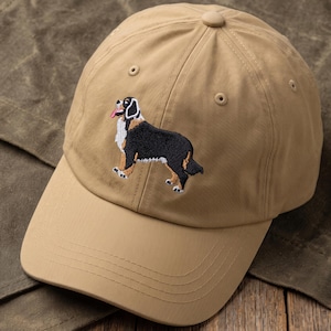 May include: A tan baseball cap featuring an embroidered Bernese Mountain Dog on the front panel. The dog is depicted in black, white, and brown thread with its tongue out. The cap has a curved brim, a button on top, and stitched eyelets.