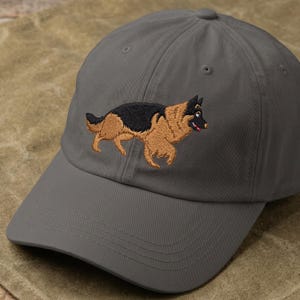 May include: Gray baseball cap featuring an embroidered German Shepherd dog in black and tan. The dog is depicted in a running pose. The cap has a curved brim and a fabric strap closure.