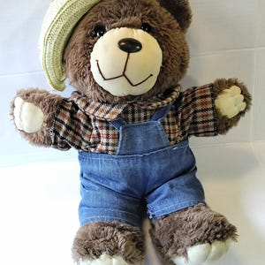 May include: A brown teddy bear wearing a plaid shirt, blue denim overalls, and a yellow straw hat. The bear has a cream-colored face, paws, and feet. The bear's fur is a dark brown color.