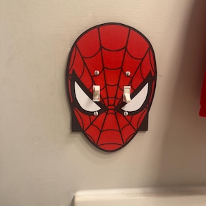 May include: A red and black Spider-Man mask shaped double light switch wall plate cover. The design features a classic web pattern and white eye shapes, with two white toggle switches protruding through the centre of the mask face.