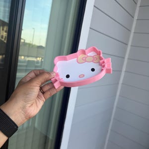 Könnte beinhalten: Ein rosa-weißes Hello Kitty-förmiges Tablett. Das Tablett hat einen rosa Umriss, einen weißen Innenraum und schwarze Augen. Eine gelbe Schleife befindet sich oben auf dem Tablett. Das Tablett wird von einer Person gehalten.