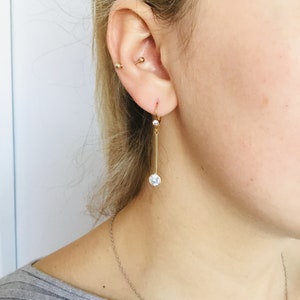 May include: Gold dangle earrings with a small clear crystal at the top and a larger clear crystal at the bottom.