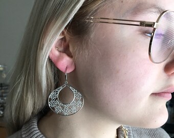 May include: Silver filigree earrings with a teardrop shape and a circular design. The earrings are hanging from a person's ear.