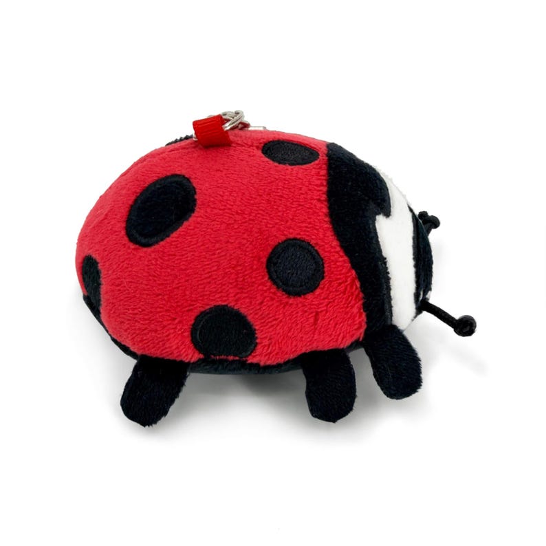 Peut inclure: Un jouet coccinelle en peluche avec un corps rouge et des taches noires. La coccinelle a des ailes et des pattes noires, et une petite boucle rouge pour l'attacher &agrave; un porte-cl&eacute;s. Le jouet est sur fond blanc.