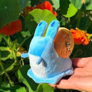 May include: A plush snail toy with a light blue body, white underbelly, and orange shell. The snail has two blue antennae and a silver keychain clip. The background features green leaves and orange flowers.