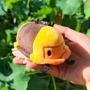 May include: A plush toy shaped like a yellow and brown insect with a small black antenna. The toy has a brown loop and a silver clasp for attaching to a keychain. The toy is held in a hand against a background of green leaves.