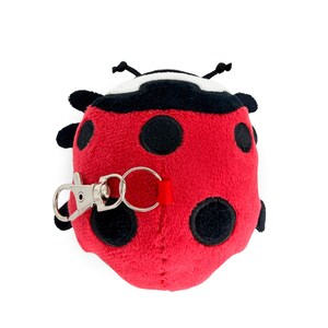 Peut inclure: Un porte-cl&eacute;s coccinelle en peluche rouge avec des pois noirs et une t&ecirc;te noire. Il est &eacute;quip&eacute; d'un mousqueton argent&eacute; et d'une boucle en tissu rouge pour l'attache. La coccinelle a des antennes noires.