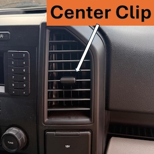 May include: A black vehicle air vent featuring a horizontal center clip, with an orange rectangular overlay containing the black text Center Clip and a white arrow pointing to the clip, positioned next to a numeric keypad and a 12V power outlet.
