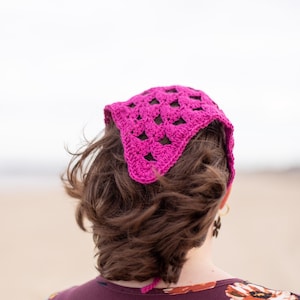 Puede incluir: Un pañuelo de ganchillo fucsia brillante con un diseño geométrico calado. El cubrecabezas es triangular y se ata en la parte posterior. La persona lleva una blusa con estampado floral. El fondo es una escena de playa borrosa.