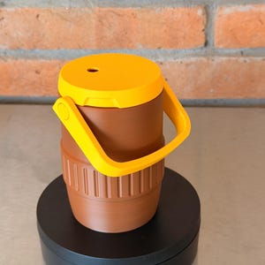 May include: A brown and yellow insulated tumbler with a handle. The tumbler has a yellow lid with a small hole and a yellow handle. The body of the tumbler is brown with vertical ridges near the bottom.