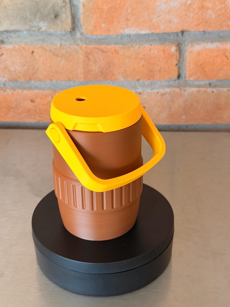 May include: A brown and yellow insulated tumbler with a handle. The tumbler has a yellow lid with a small hole and a yellow handle. The body of the tumbler is brown with vertical ridges near the bottom.