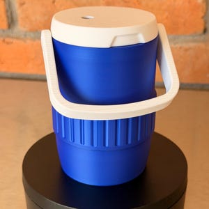 May include: A blue and white cooler with a white handle. The cooler is cylindrical with a white lid and a blue body. The handle is white and arches over the top. The cooler is sitting on a black surface.