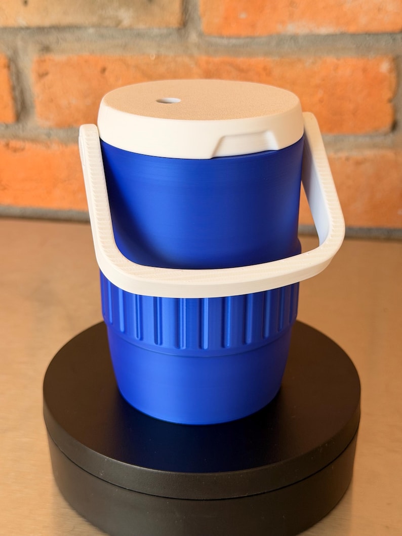 May include: A blue and white cooler with a white handle. The cooler is cylindrical with a white lid and a blue body. The handle is white and arches over the top. The cooler is sitting on a black surface.