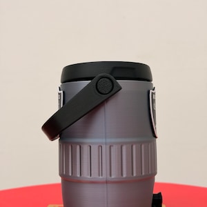 May include: A grey and black water cooler with a black handle and a black spout. The cooler has a cylindrical shape with vertical ridges around the middle. Two small black and white labels are on the sides.