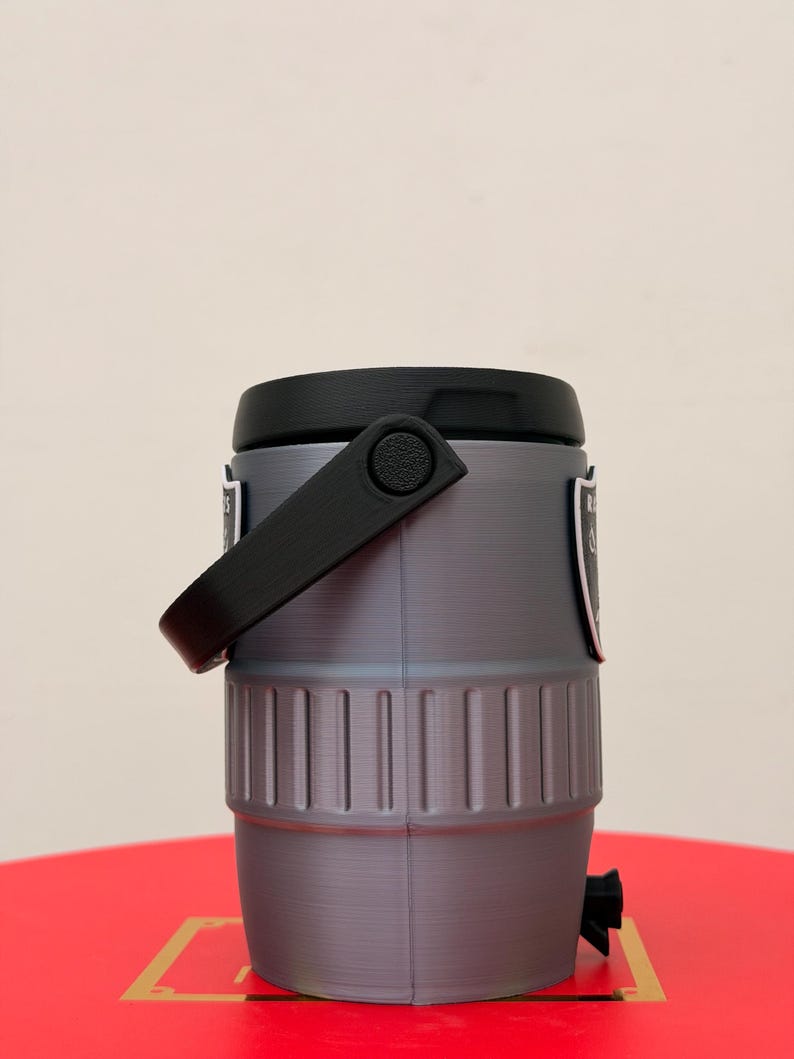 May include: A grey and black water cooler with a black handle and a black spout. The cooler has a cylindrical shape with vertical ridges around the middle. Two small black and white labels are on the sides.