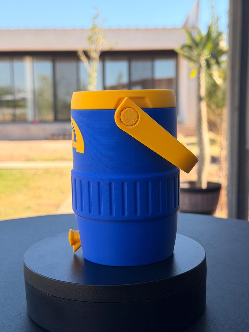 May include: A blue and yellow water cooler with a yellow handle and lid. The cooler has a spigot and a logo on the side. The cooler is sitting on a black surface.