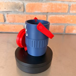 May include: A small, navy blue bucket with a red handle and accents. The bucket has a ridged design around the base and a white graphic on the side. The bucket is sitting on a black, round base.