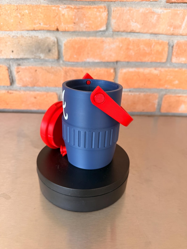 May include: A small, navy blue bucket with a red handle and accents. The bucket has a ridged design around the base and a white graphic on the side. The bucket is sitting on a black, round base.