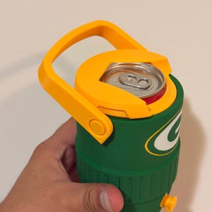 Puede incluir: Nevera aislante verde y amarilla con asa. La nevera tiene tapa y asa amarillas, y cuerpo verde con logo blanco y verde. Una lata de refresco est&aacute; dentro.