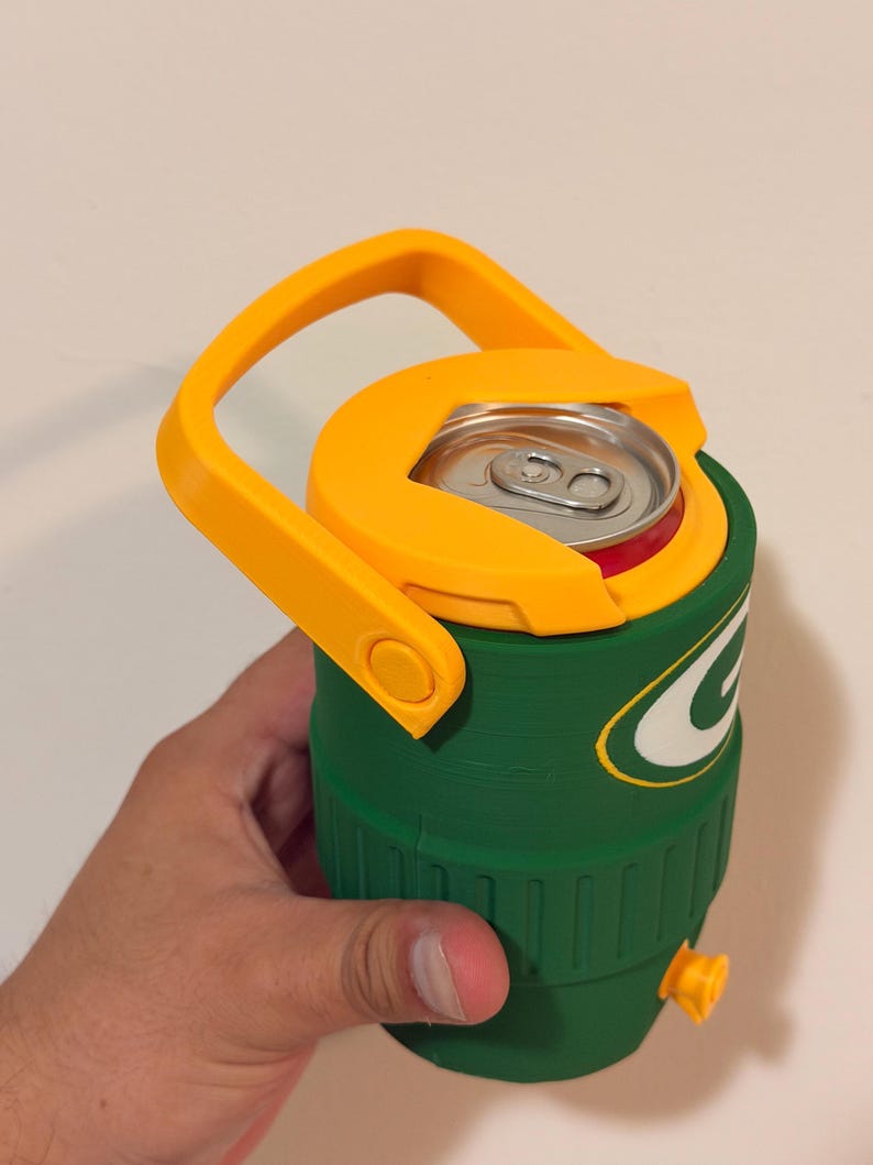 Puede incluir: Nevera aislante verde y amarilla con asa. La nevera tiene tapa y asa amarillas, y cuerpo verde con logo blanco y verde. Una lata de refresco est&aacute; dentro.