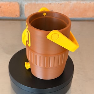 May include: A brown bucket with yellow handles and accents. The bucket has a cylindrical shape with vertical grooves near the base. The handles are attached to the sides of the bucket. The bucket is sitting on a black surface.