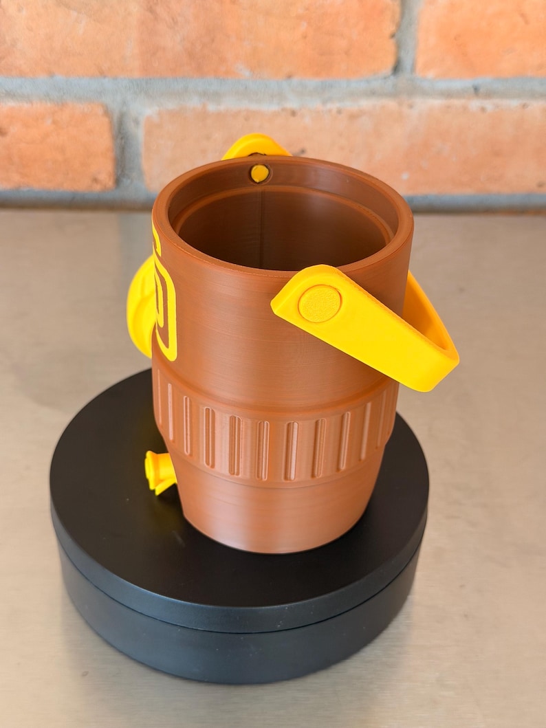 May include: A brown bucket with yellow handles and accents. The bucket has a cylindrical shape with vertical grooves near the base. The handles are attached to the sides of the bucket. The bucket is sitting on a black surface.