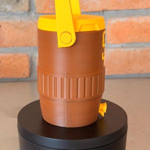 May include: A brown and yellow water cooler with a handle. The cooler has a cylindrical shape with a yellow handle and lid. The cooler has a spigot and a ribbed design around the base. The number "9" is visible on the side.