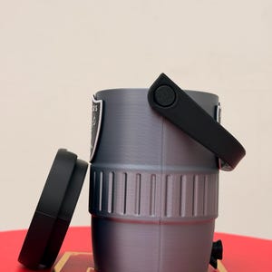 May include: A grey, cylindrical container with a black handle and lid. The container has a logo on the side and a small black spout. The lid is off to the side. The container is sitting on a red surface.