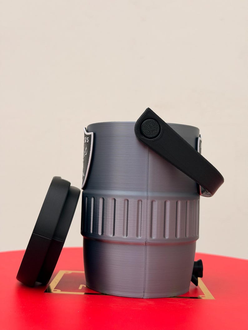 May include: A grey, cylindrical container with a black handle and lid. The container has a logo on the side and a small black spout. The lid is off to the side. The container is sitting on a red surface.