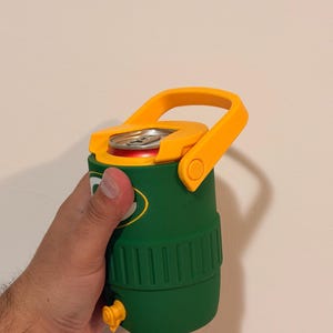 Puede incluir: Una nevera aislante verde y amarilla con asa, que contiene una lata de refresco. La nevera tiene un asa amarilla y una boquilla amarilla. La lata es roja y plateada.