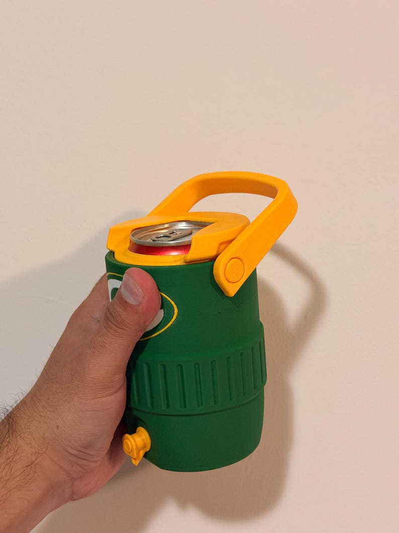 Puede incluir: Una nevera aislante verde y amarilla con asa, que contiene una lata de refresco. La nevera tiene un asa amarilla y una boquilla amarilla. La lata es roja y plateada.