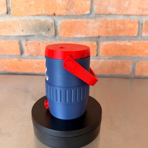 May include: A navy blue and red insulated beverage container with a handle. The container has a red lid and handle, with a white graphic on the side. The container is cylindrical with vertical grooves and a red spout at the bottom.