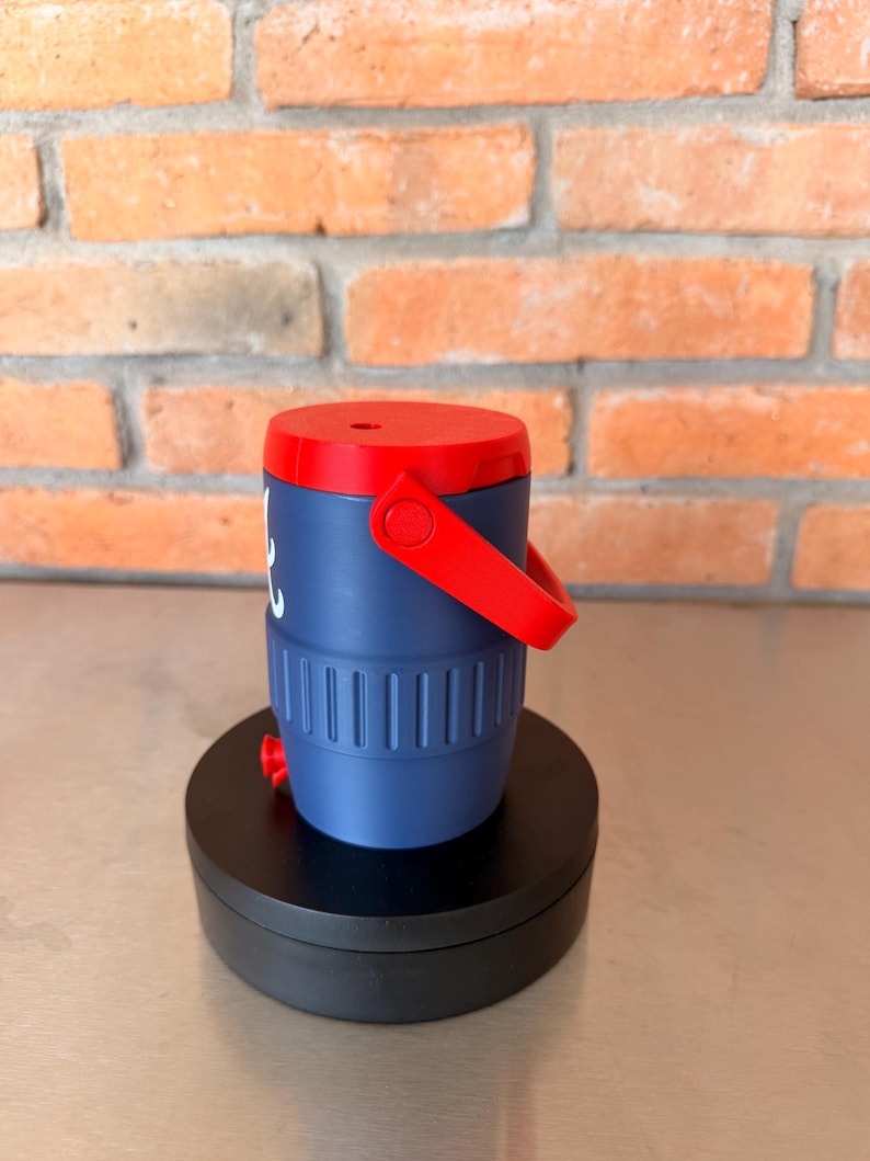 May include: A navy blue and red insulated beverage container with a handle. The container has a red lid and handle, with a white graphic on the side. The container is cylindrical with vertical grooves and a red spout at the bottom.