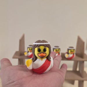 May include: A novelty rubber duck figurine with a Jesus design, featuring brown hair, a white robe with a red sash, and a yellow duck bill. Additional duck figurines are in the background, along with a small wooden structure.