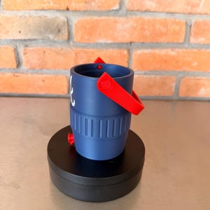May include: A navy blue bucket with a red handle and accents. The bucket has a white design on the side and is sitting on a black circular base. The background is a brick wall.