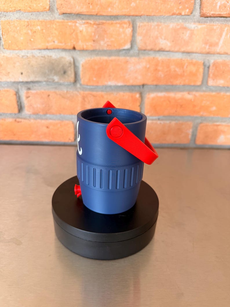May include: A navy blue bucket with a red handle and accents. The bucket has a white design on the side and is sitting on a black circular base. The background is a brick wall.