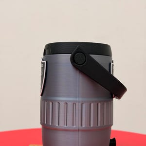 May include: A grey and black insulated water cooler with a black handle and lid. The cooler has a team logo on the side. The cooler is cylindrical with vertical ridges and a black spigot at the bottom.
