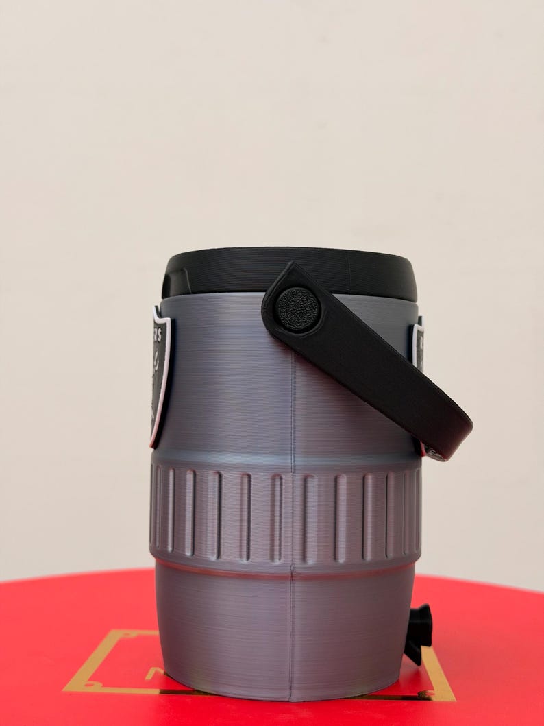 May include: A grey and black insulated water cooler with a black handle and lid. The cooler has a team logo on the side. The cooler is cylindrical with vertical ridges and a black spigot at the bottom.