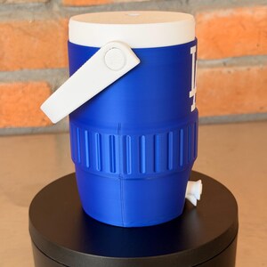 May include: A blue and white water cooler with a white handle and the Los Angeles Dodgers logo. The cooler has a white lid and a spigot. The cooler is sitting on a black surface.