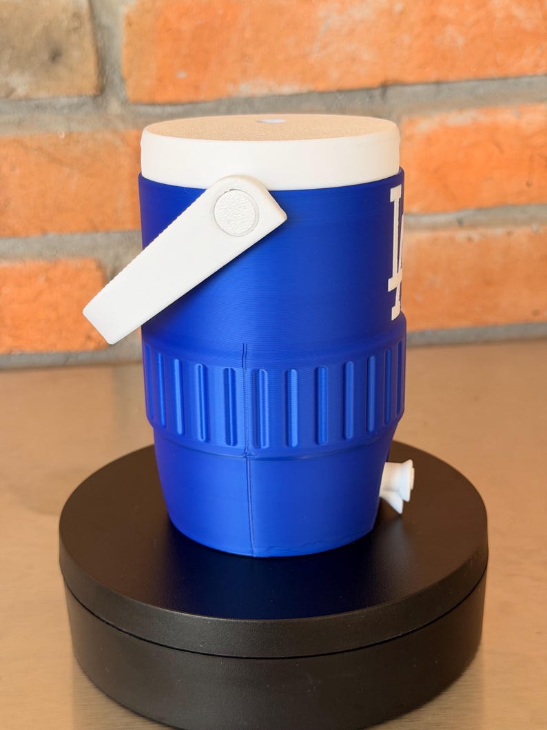 May include: A blue and white water cooler with a white handle and the Los Angeles Dodgers logo. The cooler has a white lid and a spigot. The cooler is sitting on a black surface.