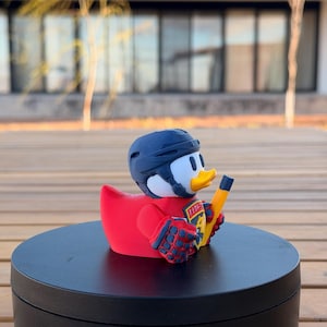 May include: A novelty rubber duck wearing a hockey helmet, red jersey, and blue gloves. The duck holds a yellow hockey stick. The jersey has the word "FISH" on it. The duck is on a black rotating stand.