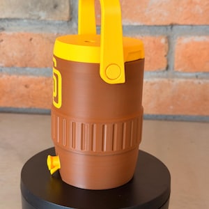 May include: Brown and yellow water cooler with a handle. The cooler has a yellow lid and handle, with a brown body. The cooler has a yellow spigot and a logo on the side. The cooler is sitting on a black surface.