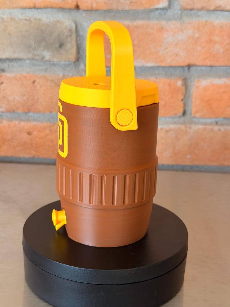 May include: Brown and yellow water cooler with a handle. The cooler has a yellow lid and handle, with a brown body. The cooler has a yellow spigot and a logo on the side. The cooler is sitting on a black surface.