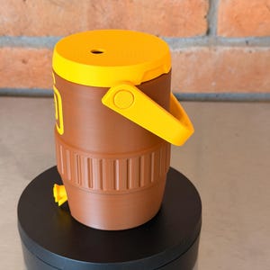 May include: A brown and yellow water cooler with a handle and a spigot. The cooler has a yellow lid and handle, and a brown body with vertical ridges. The cooler is sitting on a black rotating platform.