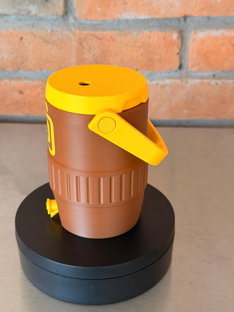 May include: A brown and yellow water cooler with a handle and a spigot. The cooler has a yellow lid and handle, and a brown body with vertical ridges. The cooler is sitting on a black rotating platform.