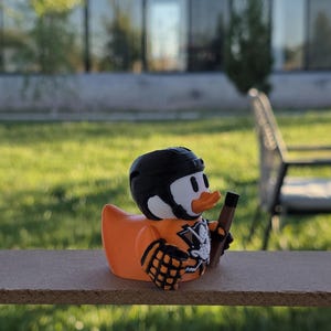Peut inclure: Un canard en plastique orange portant un casque et un maillot de hockey noirs, tenant une crosse de hockey. Le canard a un visage blanc, des yeux noirs et un bec orange. Le maillot a un motif de t&ecirc;te de mort. Le canard est sur une surface en bois.