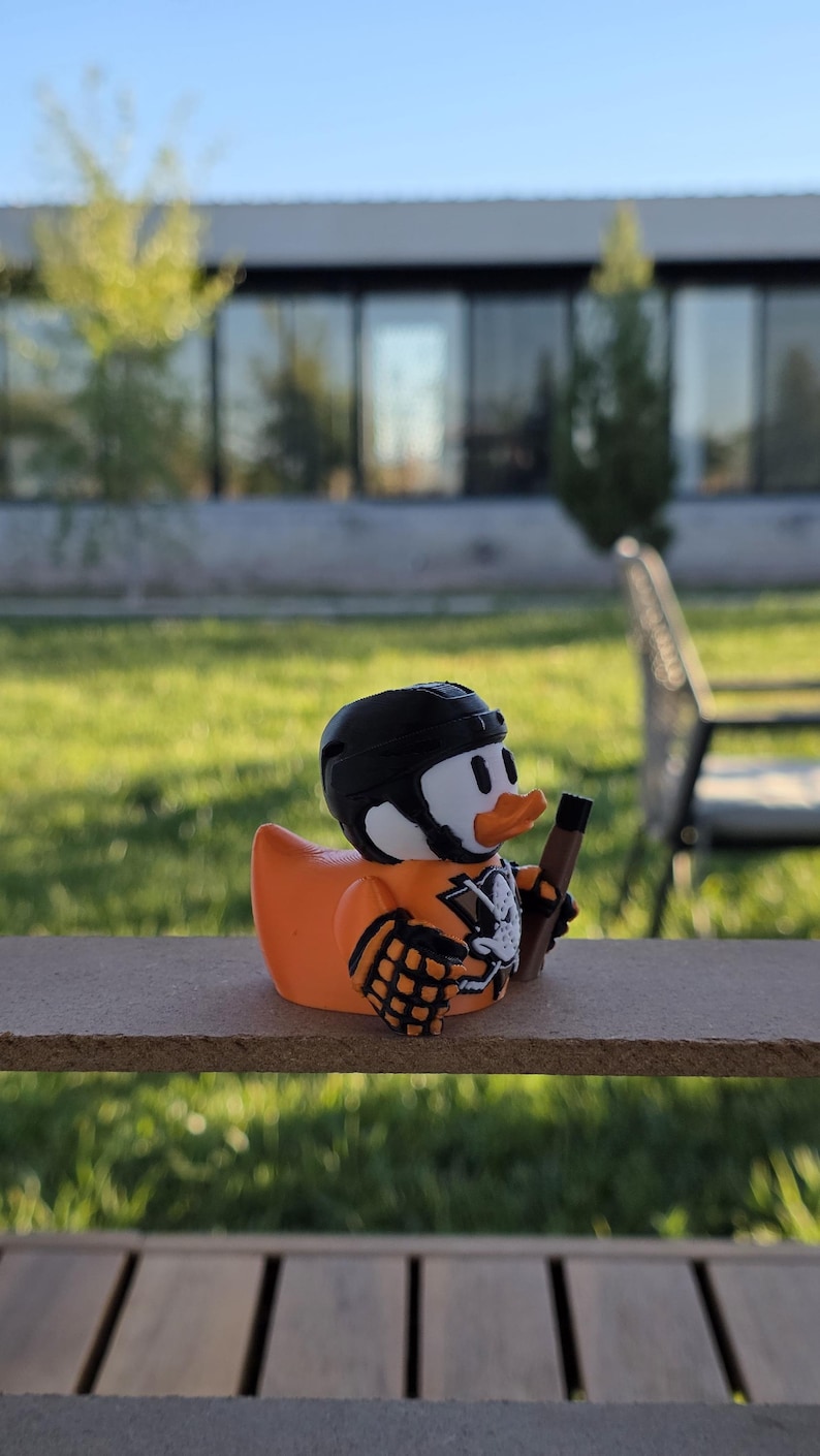 Peut inclure: Un canard en plastique orange portant un casque et un maillot de hockey noirs, tenant une crosse de hockey. Le canard a un visage blanc, des yeux noirs et un bec orange. Le maillot a un motif de t&ecirc;te de mort. Le canard est sur une surface en bois.