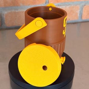 May include: A brown bucket with a yellow handle and accents, and a yellow lid. The bucket has the number "4" printed on the side in yellow. The lid has a hole in the center. The bucket is sitting on a black surface.