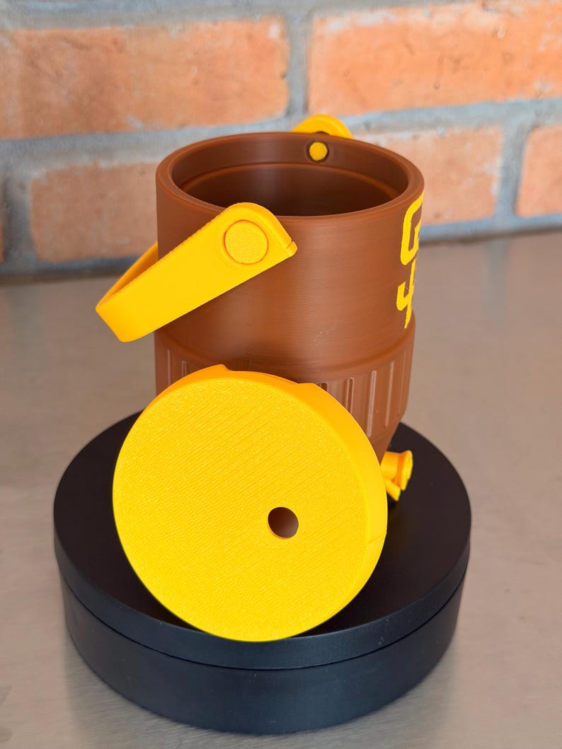 May include: A brown bucket with a yellow handle and accents, and a yellow lid. The bucket has the number "4" printed on the side in yellow. The lid has a hole in the center. The bucket is sitting on a black surface.
